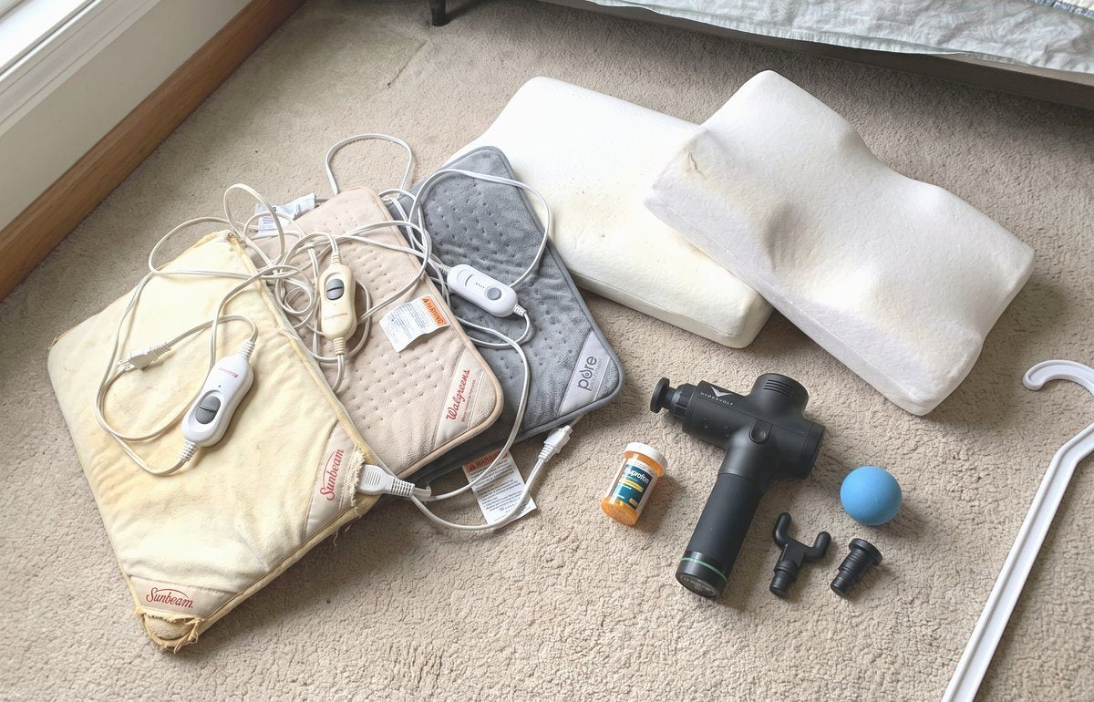 Six years of bought remedies laid out on a bedroom carpet: three heating pads of different ages, two contour pillows, a massage gun with spare heads, a lacrosse ball, a pill bottle and a long massage hook cut off by the edge of the frame.