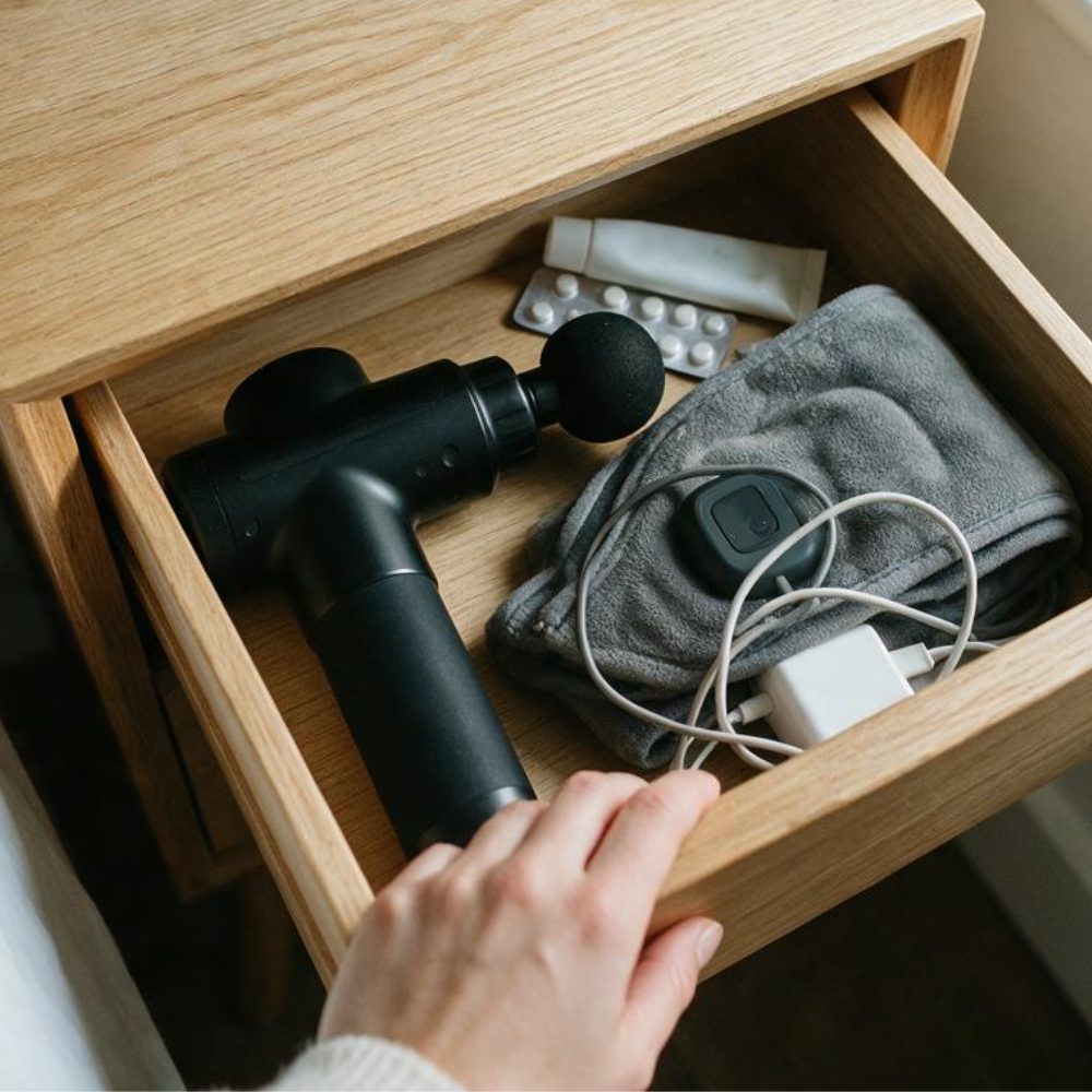 A hand opening a bedside drawer with a massage gun and a heating pad inside