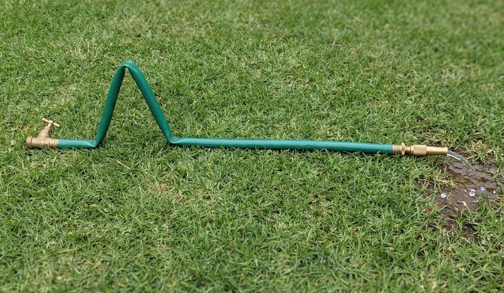 A green garden hose lying on a lawn. Next to the brass tap on the left, the hose is folded in one sharp kink. At the far end, a brass nozzle lets out only a thin dribble of water.